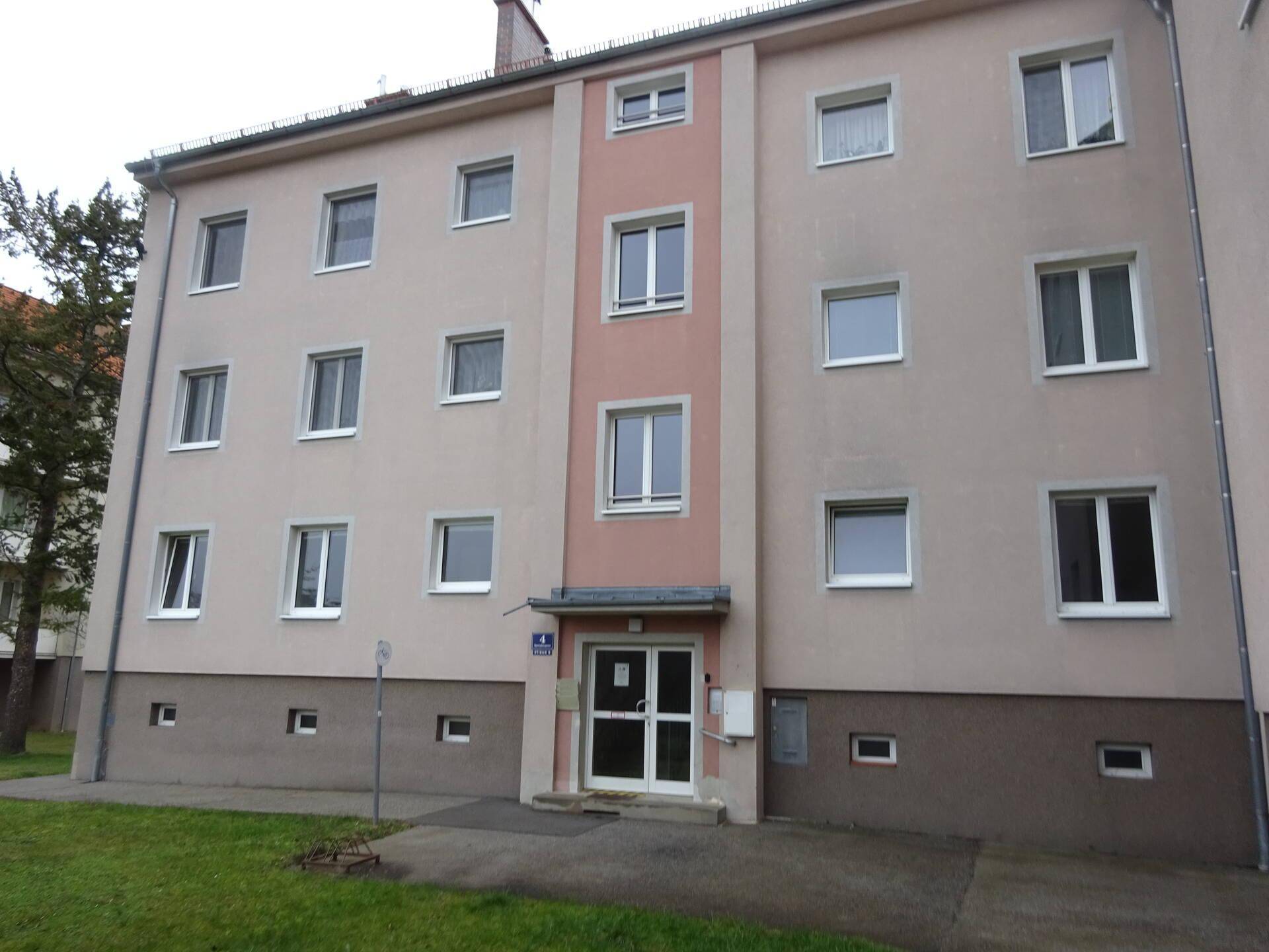 Mehrstöckiges Wohngebäude mit beigefarbener Fassade, vielen Fenstern und einem kleinen Vorgarten.