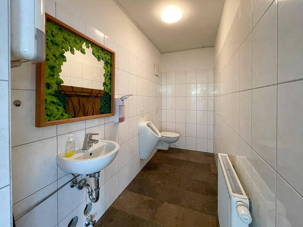 Einfache Toilette mit weißen Fliesen, Urinal und Waschbecken, ausgestattet mit einem Heizkörper.