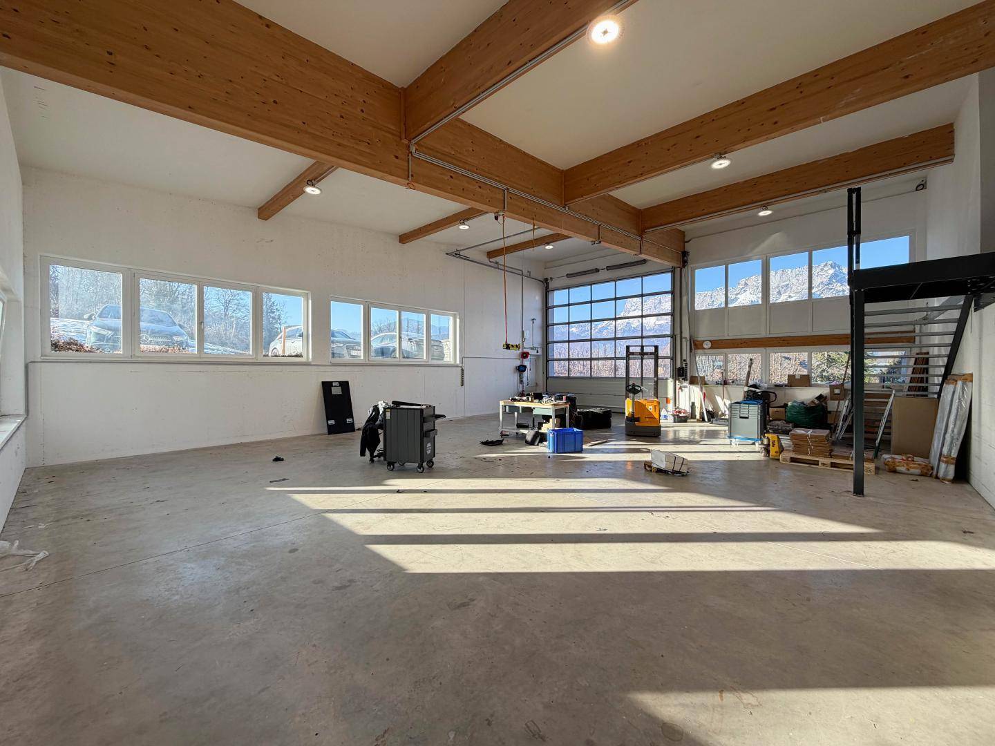 Weitläufige Halle mit Betonboden, Holzbalken und großen Fenstern, die einen Blick auf die Berge bieten.