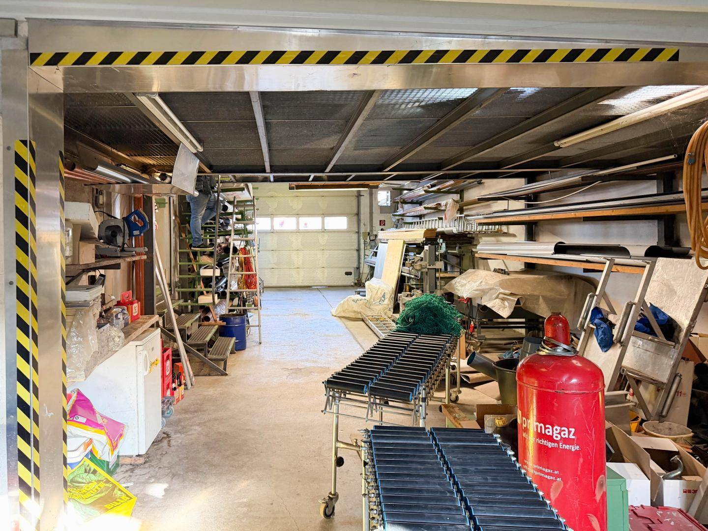 Geräumige Garage oder Lagerhalle mit Rolltor, Förderband und verschiedenen Materialien, gut beleuchtet.