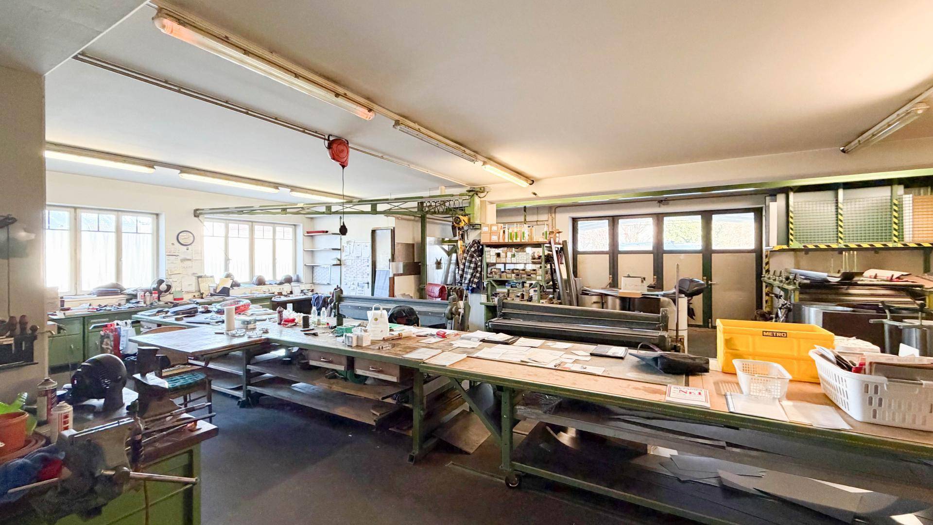 Geräumiger Werkstattbereich mit langen Arbeitstischen, Maschinen und großen Fenstern, die Tageslicht hereinlassen.