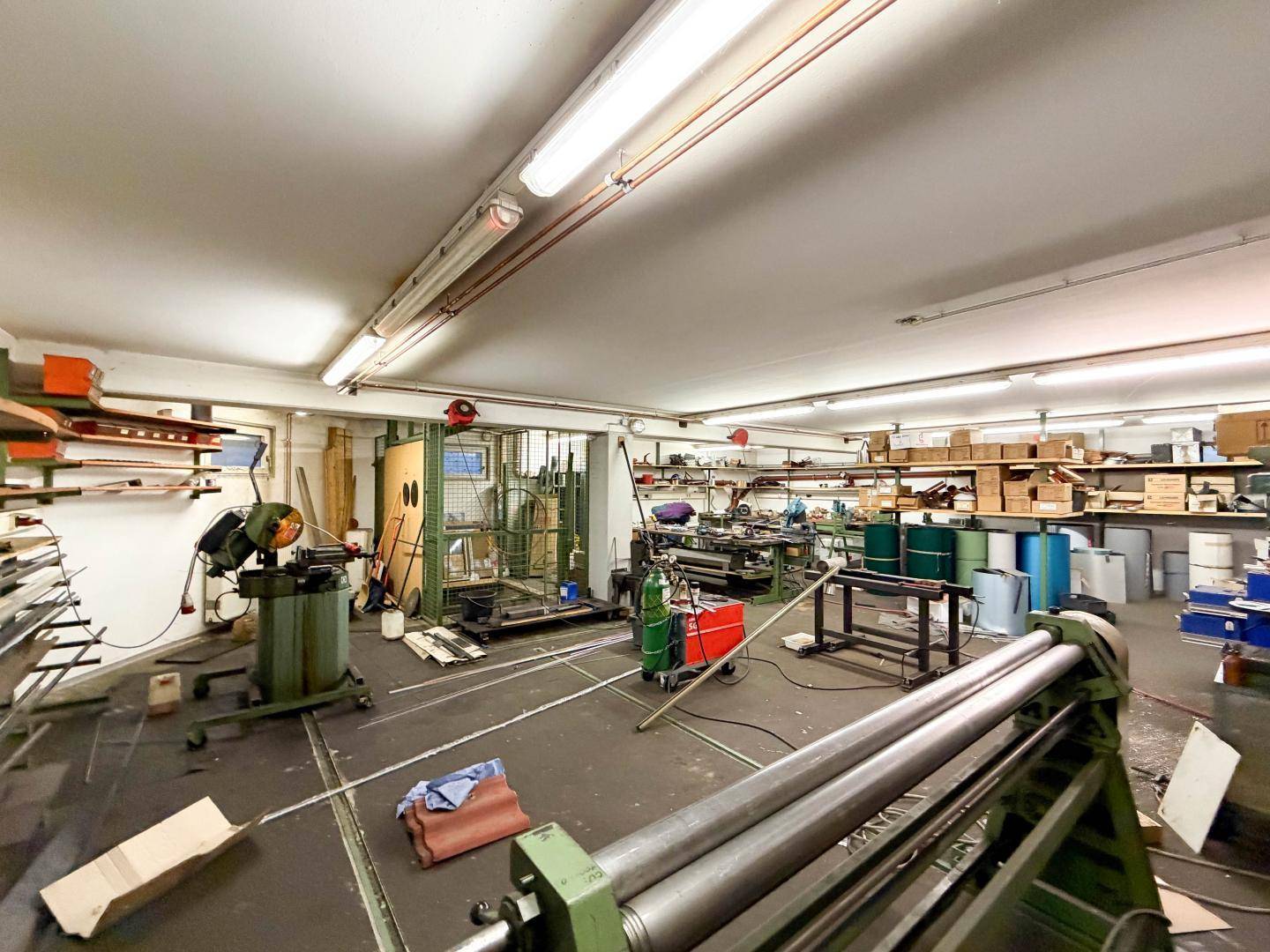 Industrieller Werkstattraum mit schweren Maschinen, Lagerregalen und verschiedenen Arbeitsbereichen, gut beleuchtet.