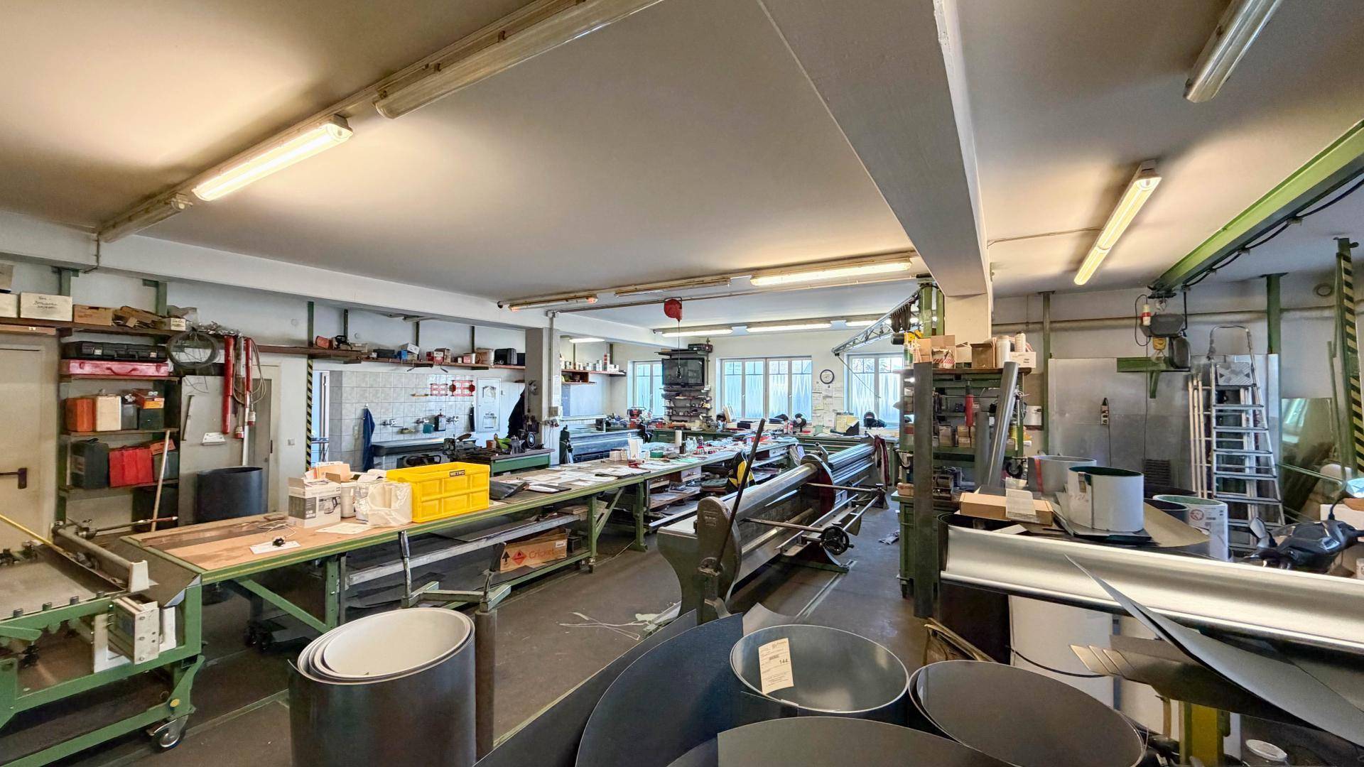 Weitläufige Werkstatt mit verschiedenen Maschinen, Werkbänken und Materialien, gut beleuchtet durch Fenster und Deckenlampen.