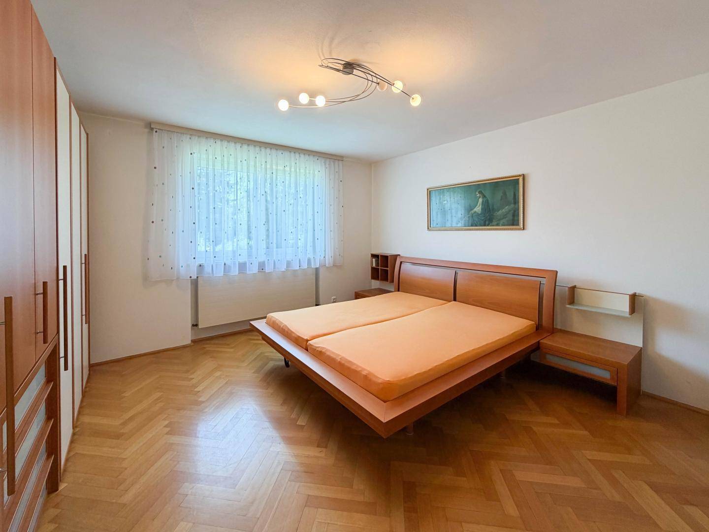 Helles Schlafzimmer mit Holzbett, Parkettboden und großem Fenster mit Vorhängen.