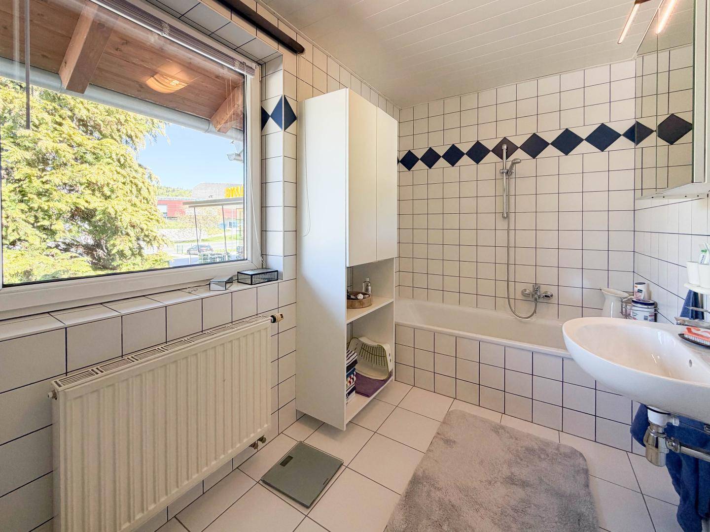 Badezimmer mit Badewanne, Waschbecken, Fenster und weißen Fliesen mit blauen Akzenten.