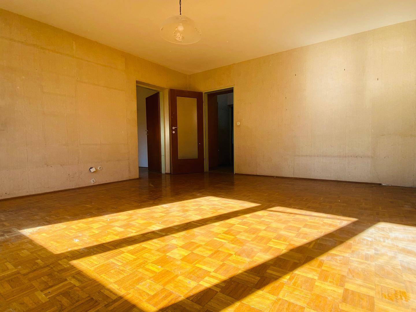 Geräumiger Wohnraum mit Parkettboden und zwei Türen, durchflutet von natürlichem Licht.