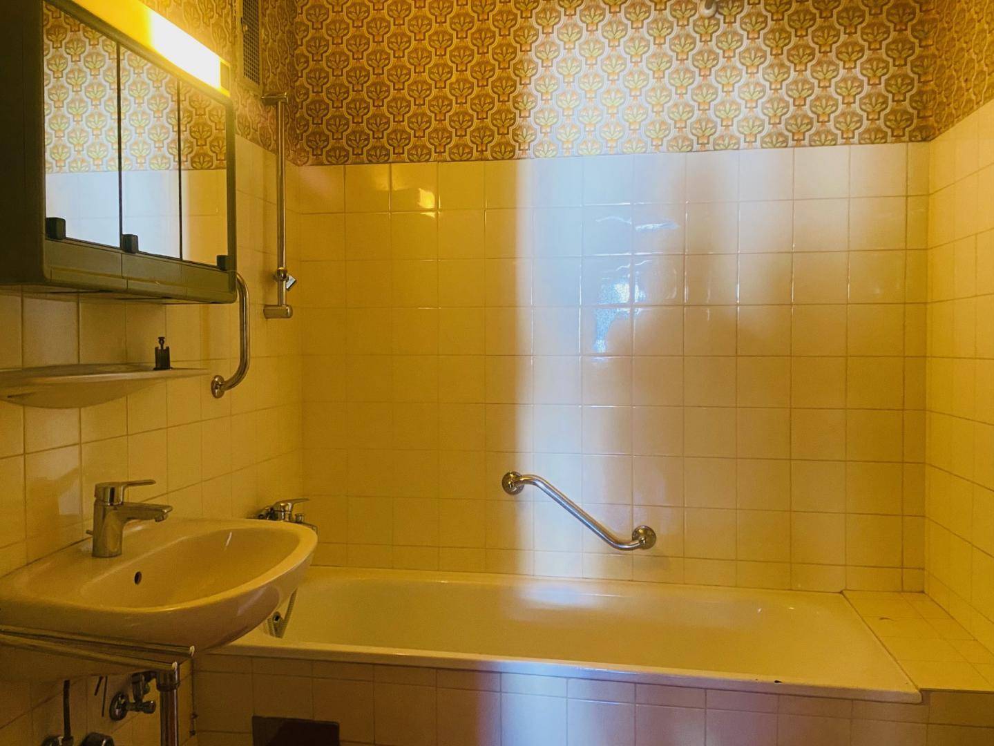 Badezimmer mit Badewanne, Waschbecken und Fliesen, teilweise mit gemusterter Tapete.