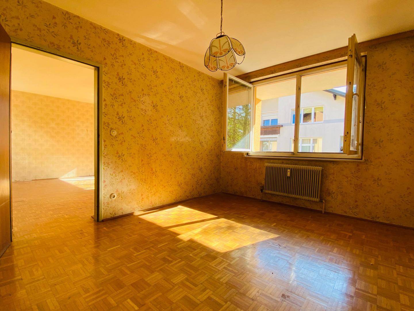 Sonniger Wohnbereich mit Parkettboden, älterer Tapete und Zugang zu einem weiteren Zimmer.