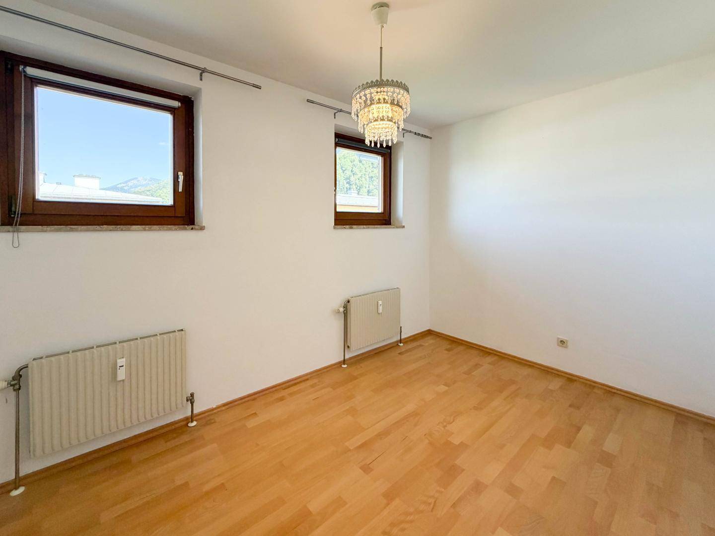 Helles Schlafzimmer mit Parkettboden, zwei Fenstern und einem Kronleuchter.