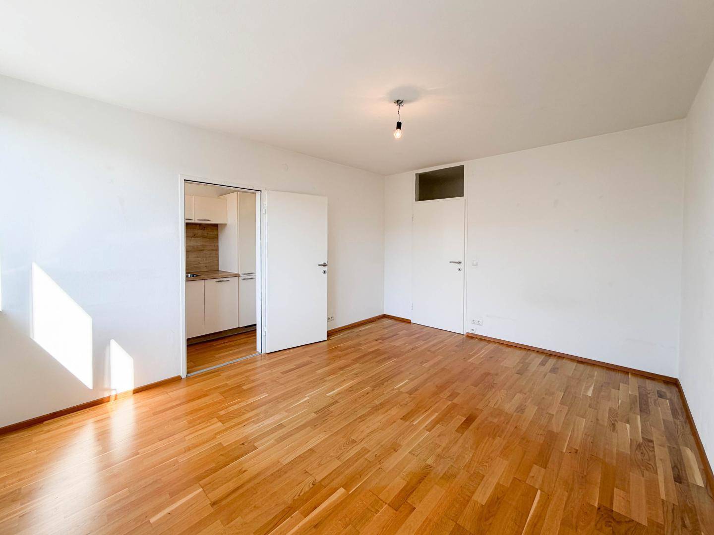 Geräumiges Wohnzimmer mit Holzboden und offenem Zugang zu einer kleinen Küche.
