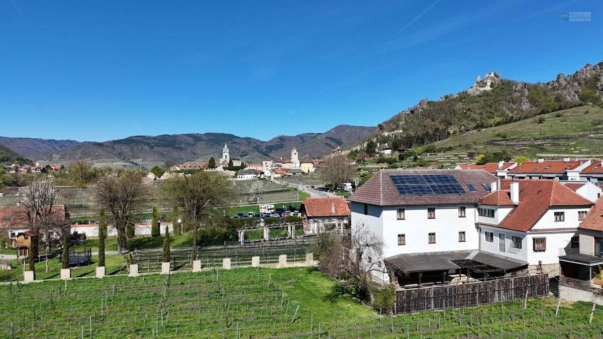 Luftaufnahme eines Gebäudes mit Solaranlage auf dem Dach, umgeben von Weinbergen und einem Dorf im Hintergrund.