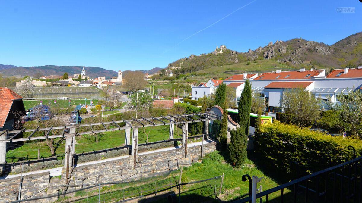 Weitläufiger Blick von einer Terrasse auf grüne Weinberge, ein Dorf und eine Burgruine in der Ferne.
