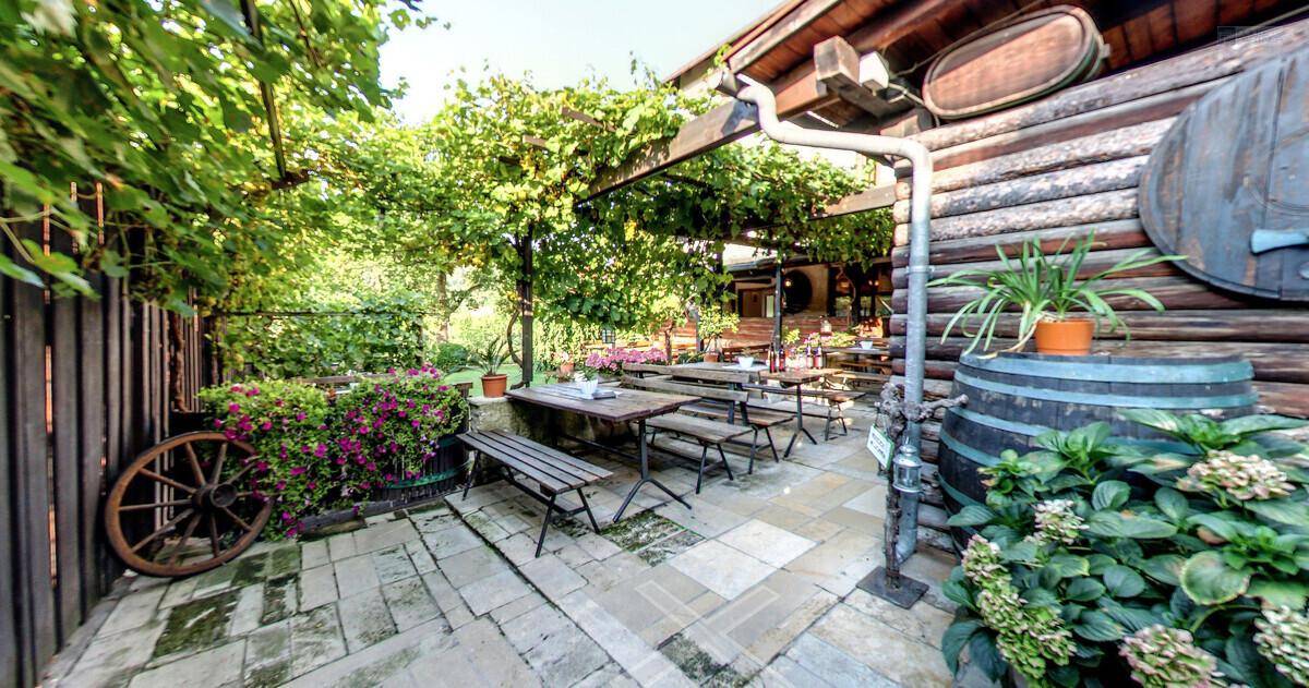 Rustikaler Außenbereich mit Holztischen, Bänken und Weinfässern als Dekoration unter einer Pergola.