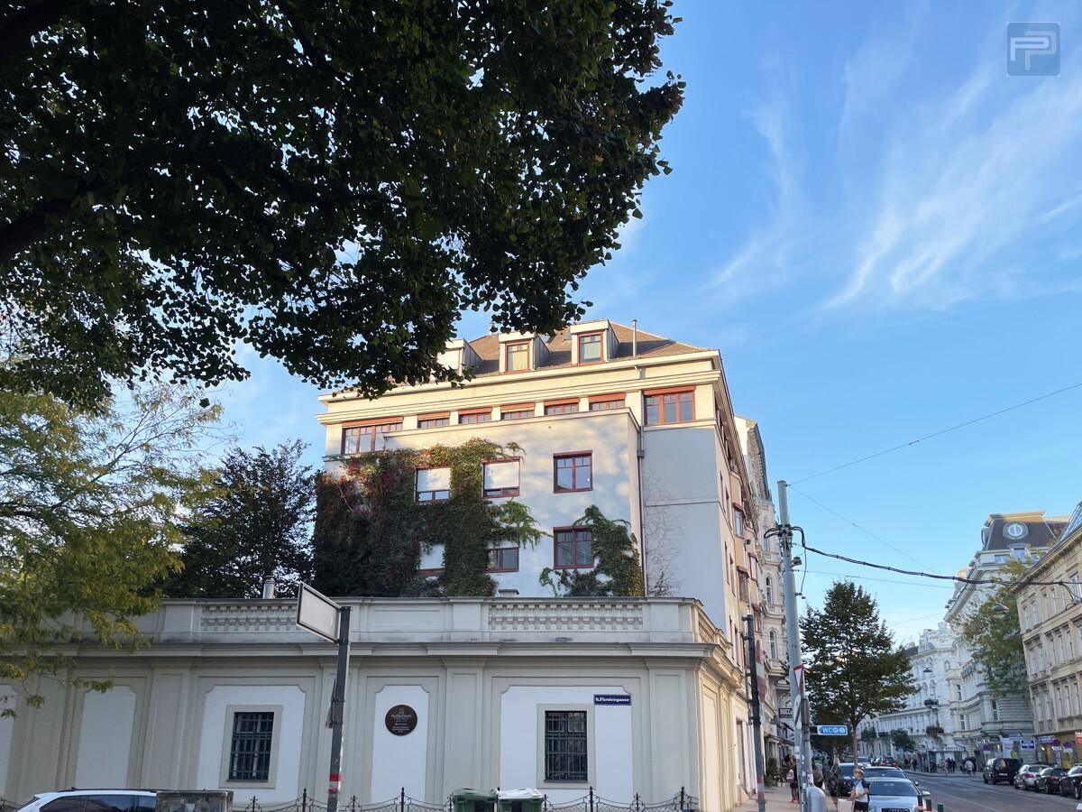 Gebäudeansicht von der Straße mit Bäumen und einem Teil der Fassade unter blauem Himmel.
