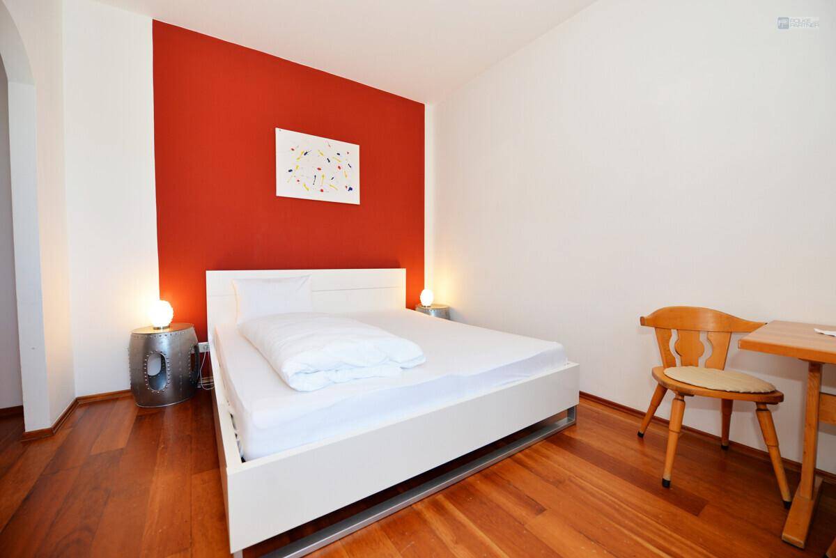 Modernes Schlafzimmer mit weißem Doppelbett, roter Akzentwand und glänzendem Holzfußboden.