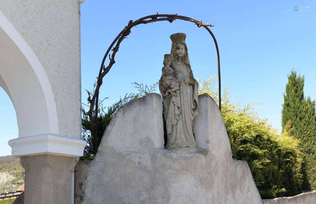 Eine Marienstatue mit Kind unter einem Bogen, umgeben von Bäumen und Hügeln.