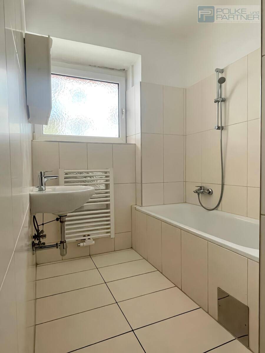 Helles Badezimmer mit weißen Fliesen, Badewanne mit Duschfunktion und einem Fenster für natürliche Belüftung.