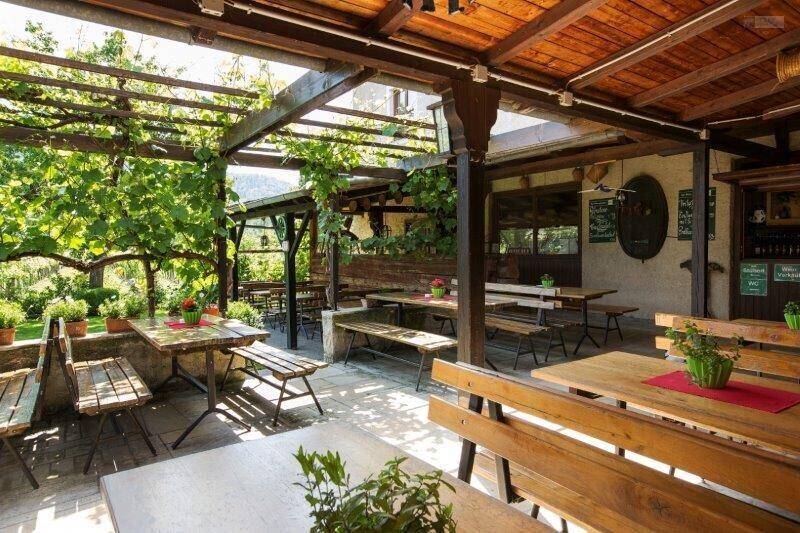Weitläufiger Außenbereich mit Holztischen und Bänken unter einer grünen Pergola, umgeben von Pflanzen.