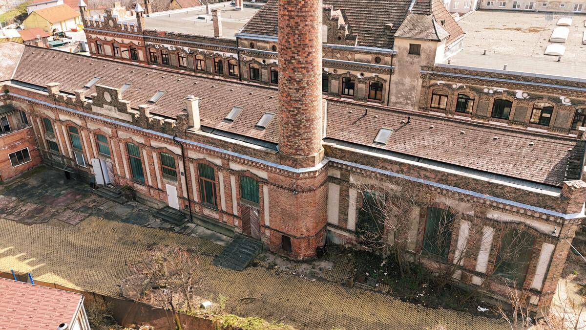 Luftaufnahme eines weitläufigen Backsteingebäudes mit hohem Schornstein und vielen Fenstern.