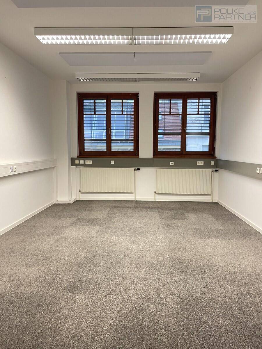 Schmalerer Blick in den leeren Büroraum mit zwei Fenstern und modernen Deckenleuchten.