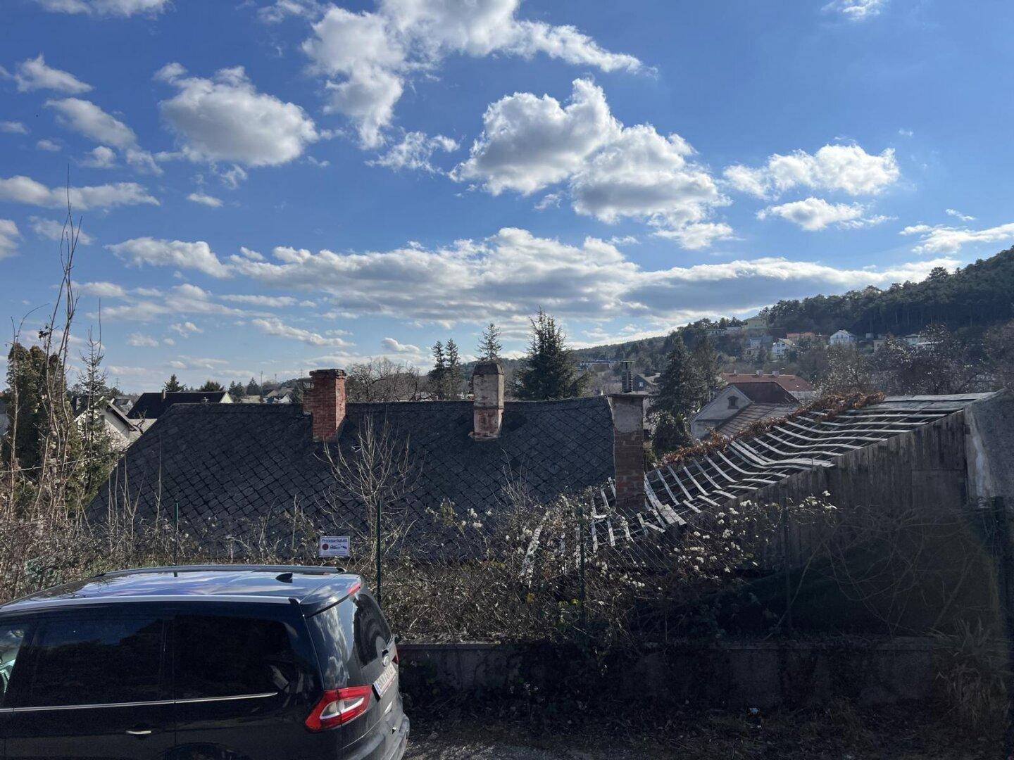 Blick auf alte Gebäude mit Dacheinsturz und verwildertem Grundstück, mit einem schwarzen Auto im Vordergrund.