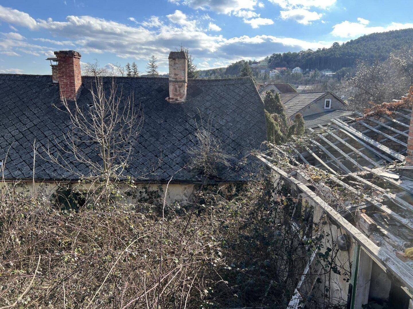Altes Haus mit Ziegeldach und Schornsteinen neben einem Gebäude mit eingestürztem Dachstuhl und freiliegenden Holzbalken.