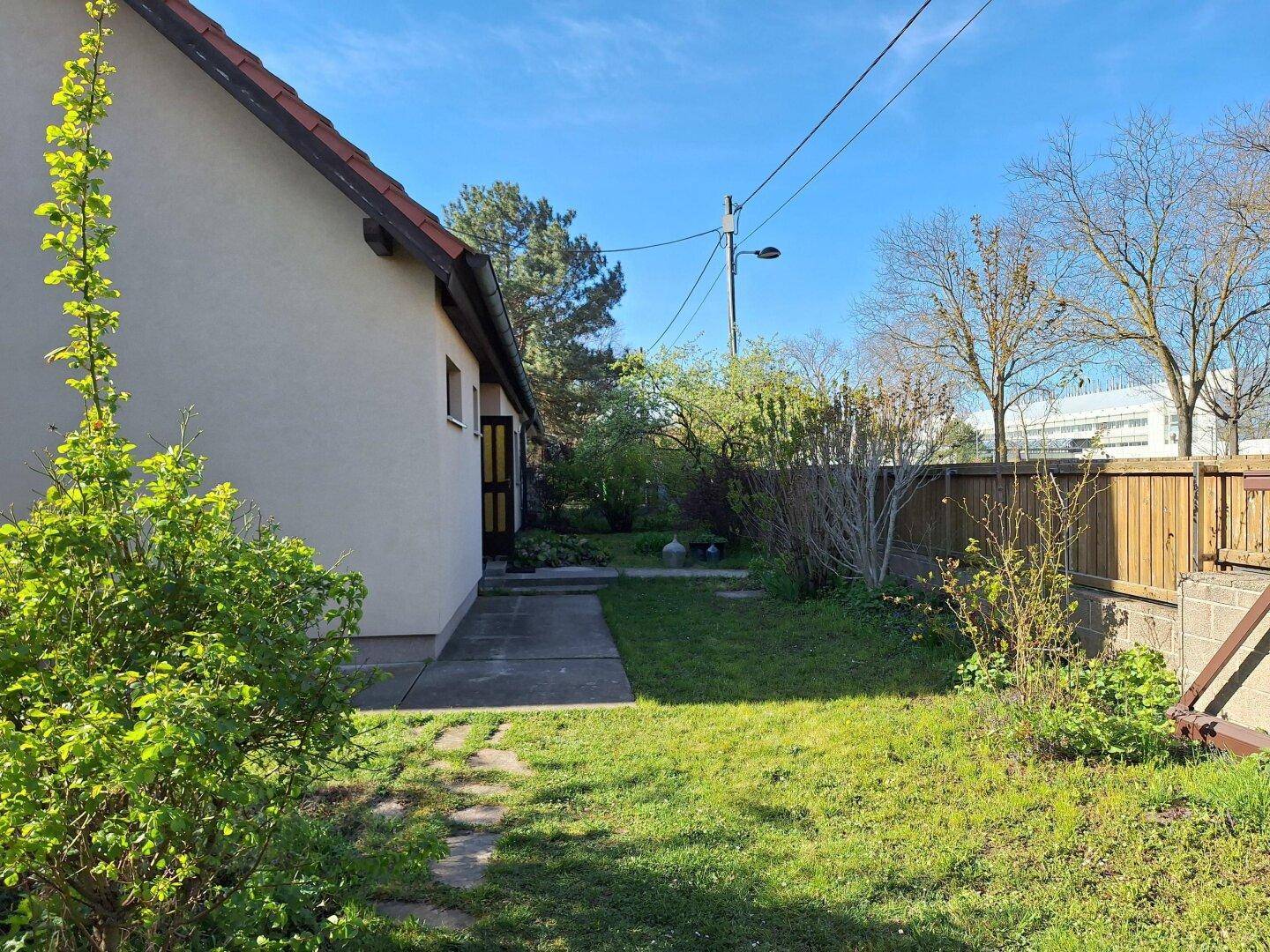 Seitenansicht des Hauses mit einem Gartenweg und grüner Bepflanzung unter blauem Himmel.