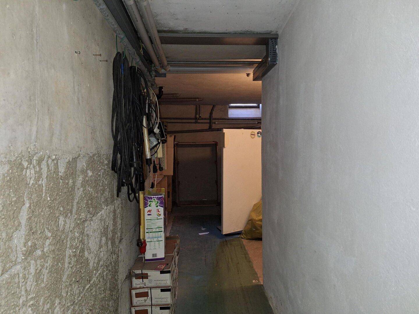 Schmaler Kellerflur mit sichtbaren Leitungen und Kabeln, der zu weiteren Kellerräumen führt.