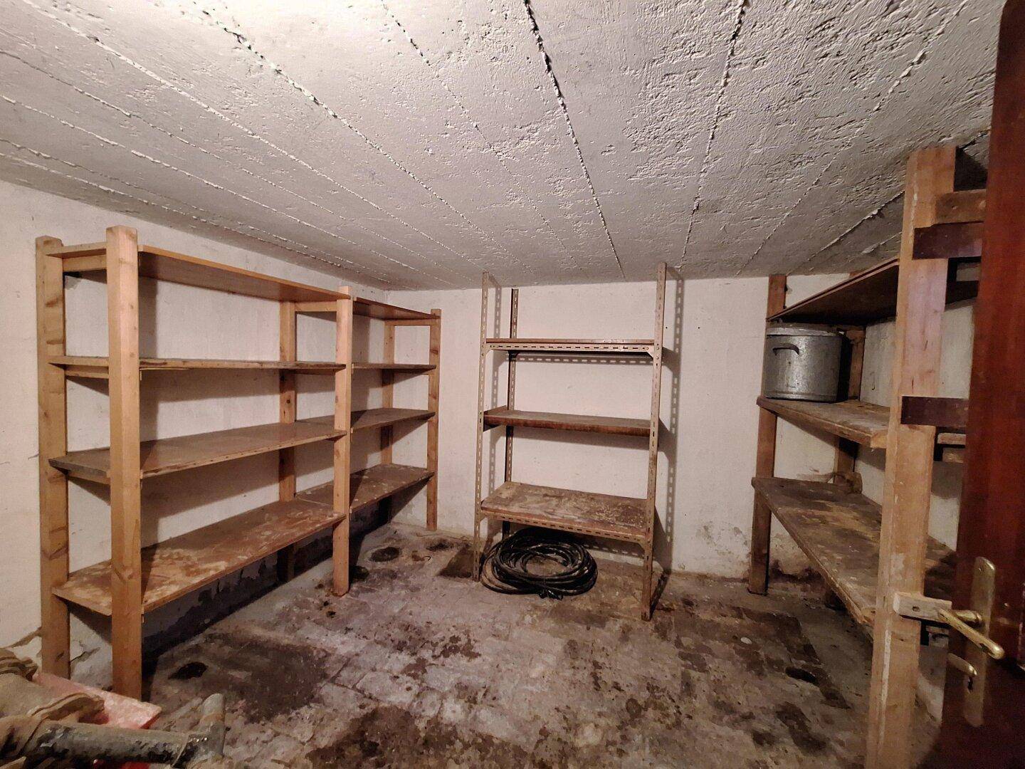 Kleinerer Kellerraum mit Holz- und Metallregalen, unverputzter Decke und schmutzigem Boden.