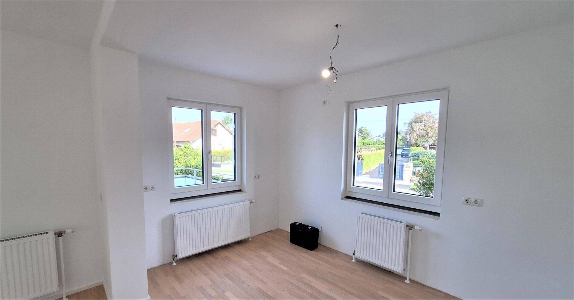 Helles Zimmer mit Parkettboden und zwei Fenstern, die viel Tageslicht hereinlassen.