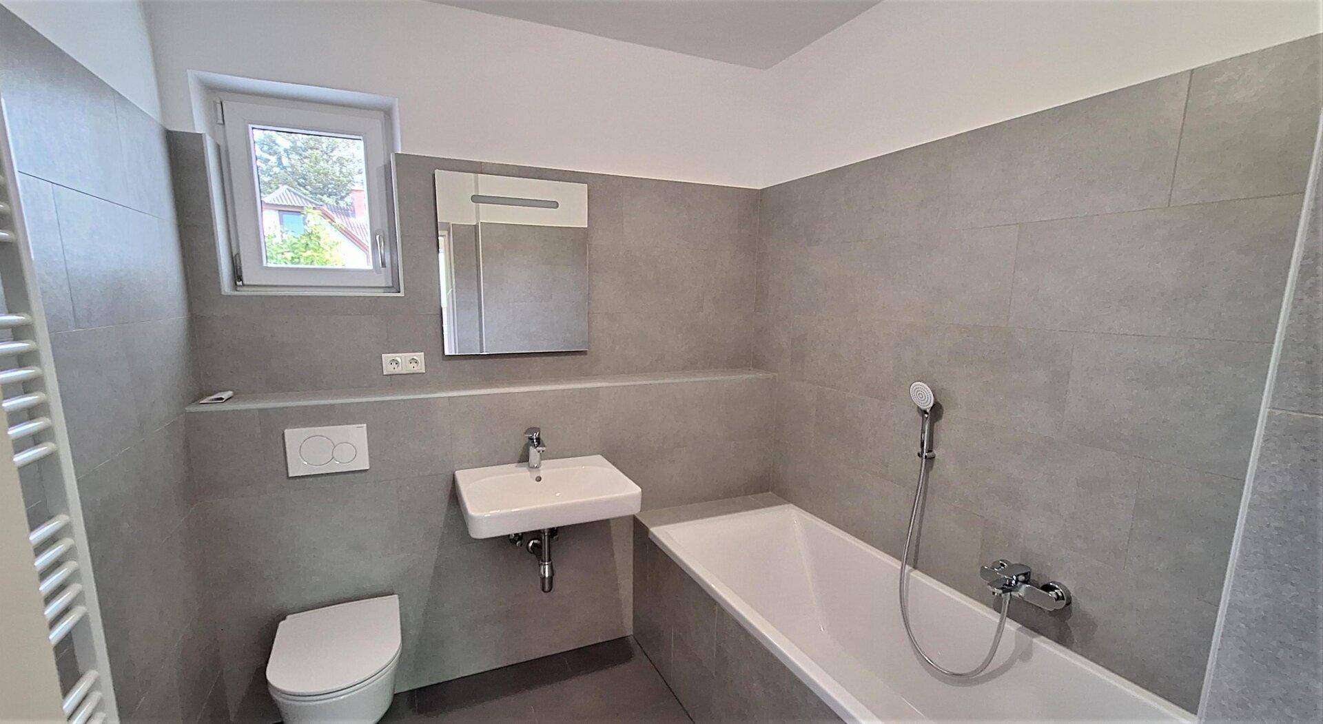 Modernes Badezimmer mit grauen Fliesen, Badewanne, Waschbecken und Fenster für Tageslicht.