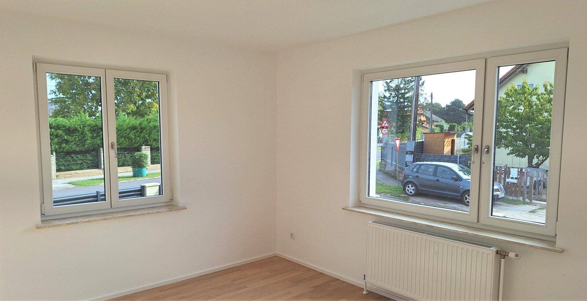 Helles Zimmer mit Parkettboden und zwei Fenstern, die einen Blick nach draußen bieten.