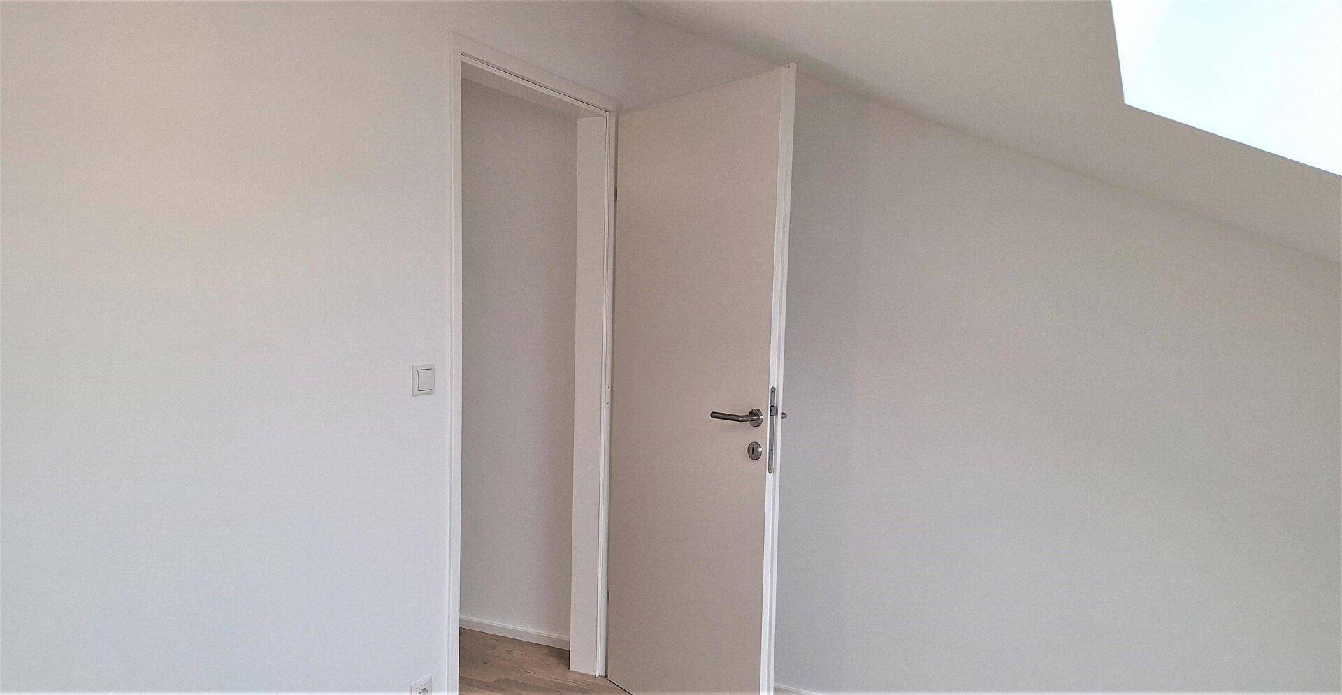 Offene weiße Tür in einem frisch renovierten Raum mit Schrägdach und hellem Laminatboden.