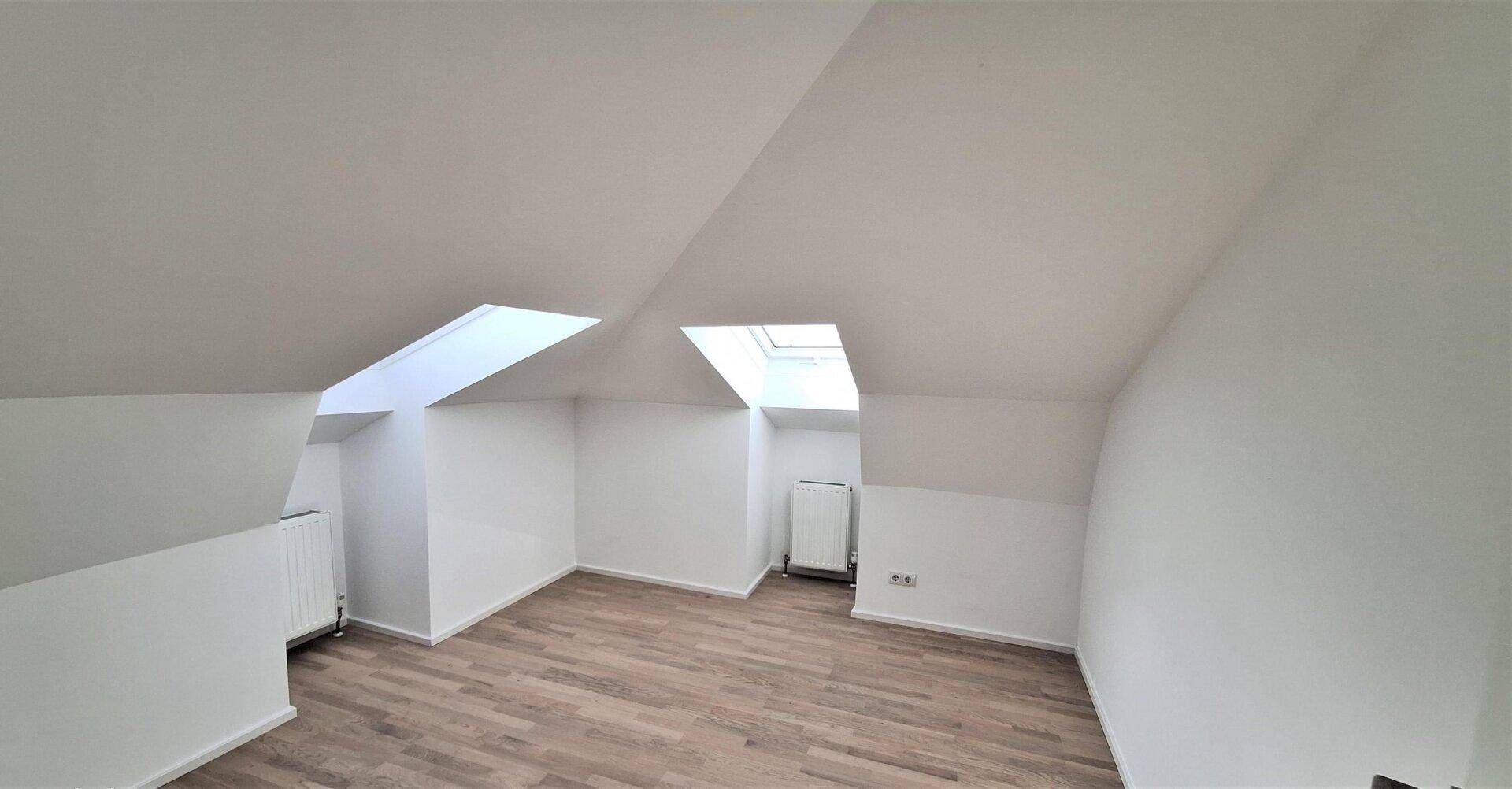 Geräumiger Dachgeschossraum mit zwei Dachfenstern, Heizkörpern und hellem Laminatboden.