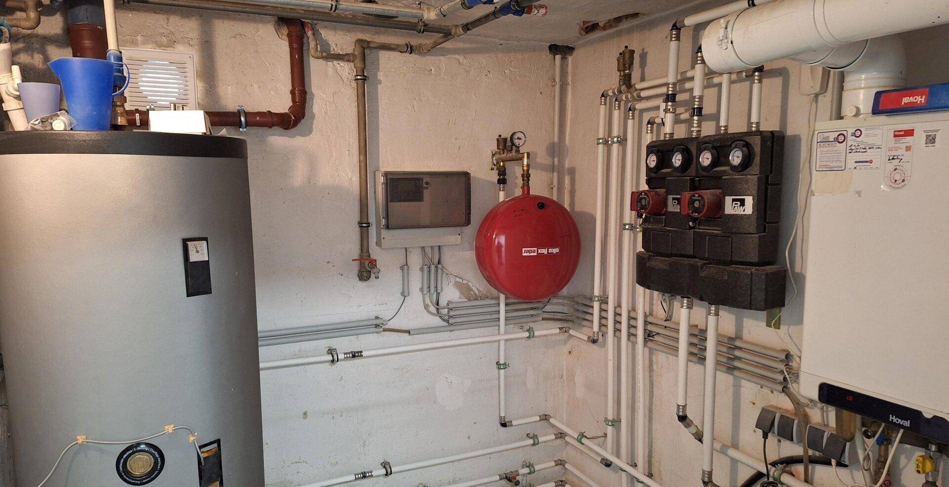 Technikraum im Keller mit Heizungsanlage, Warmwasserspeicher und zahlreichen Rohrleitungen.