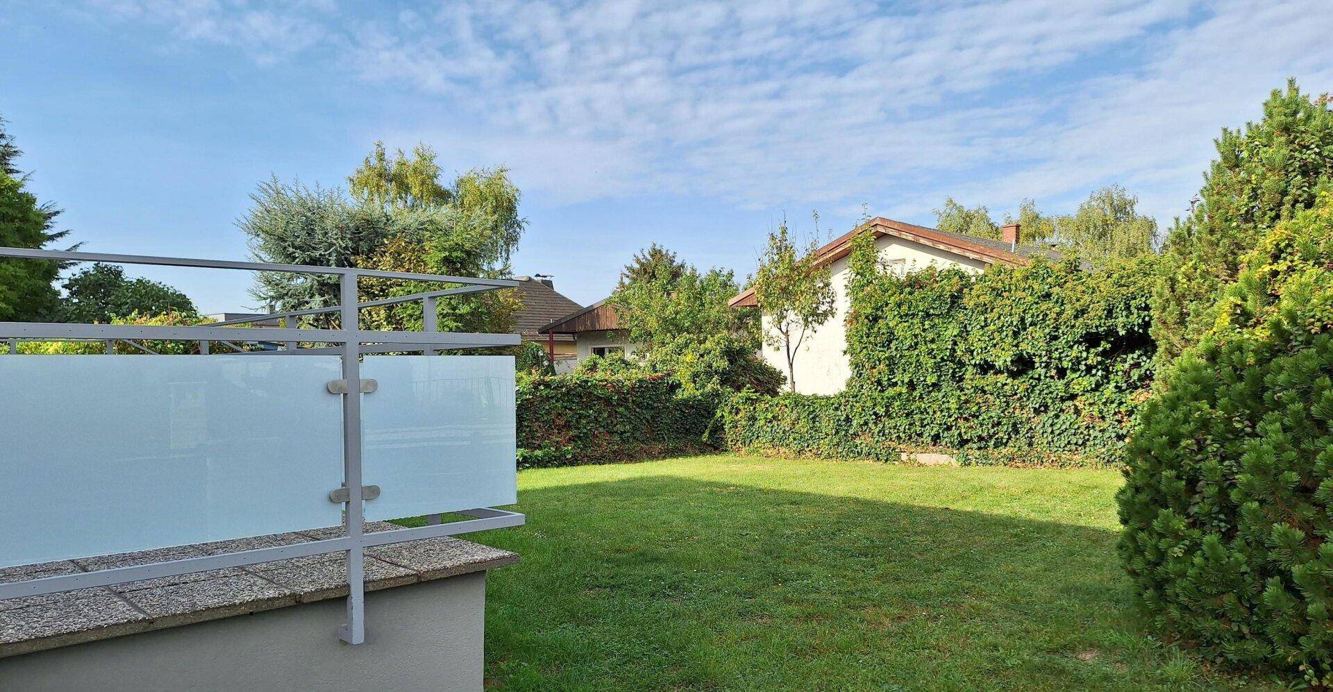 Gepflegter Garten mit Rasenfläche, Hecken und Bäumen, umgeben von einer Glasbrüstung.