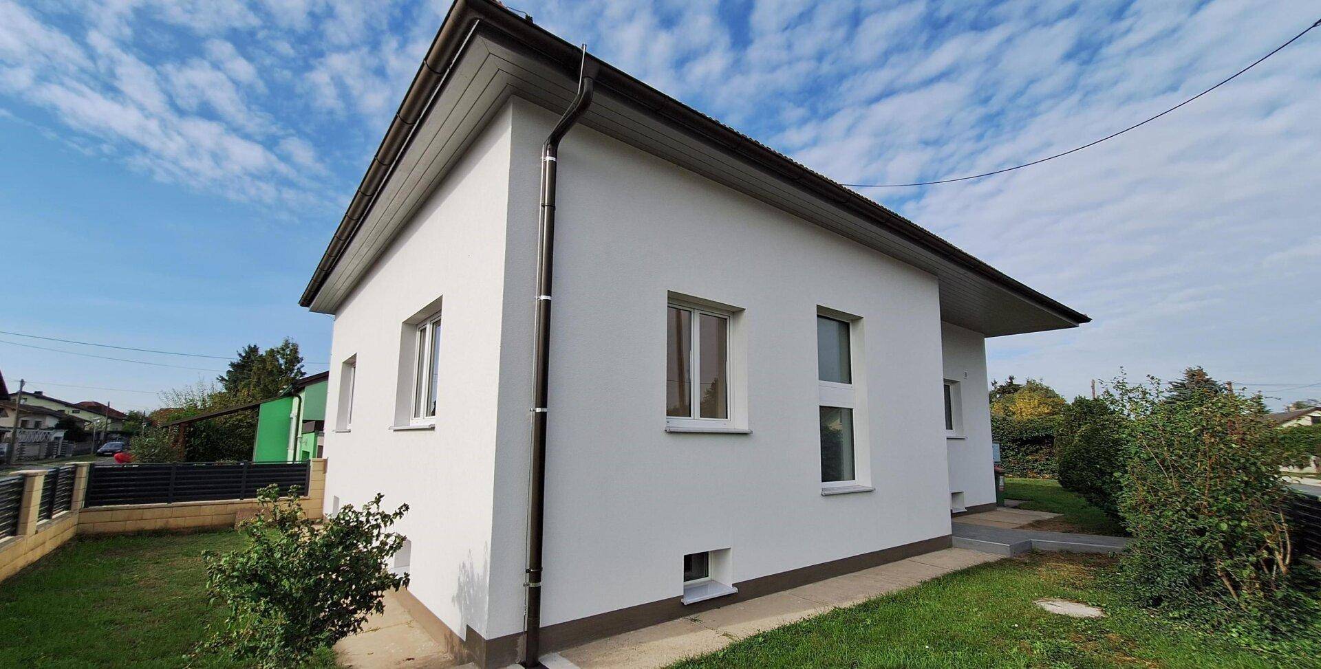 Seitenansicht des modernen Hauses mit weißer Fassade und braunem Dach, umgeben von Grün.