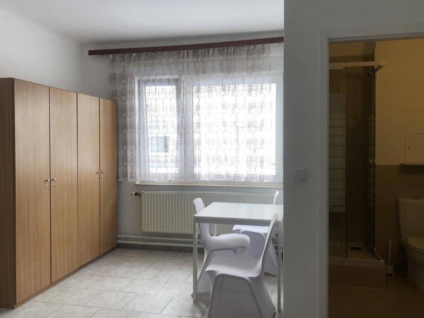 Wohnbereich mit Esstisch, Stühlen, großem Kleiderschrank und Fenster mit Vorhängen.