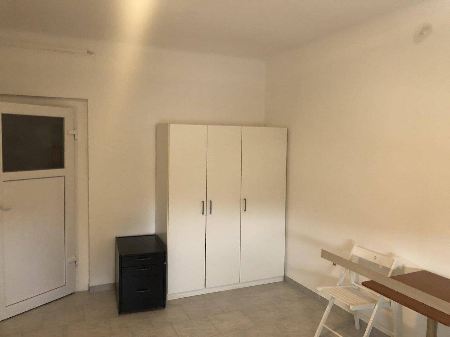 Einfacher Raum mit weißem Kleiderschrank, kleinem Tisch und Stühlen, ideal als Schlaf- oder Wohnbereich.