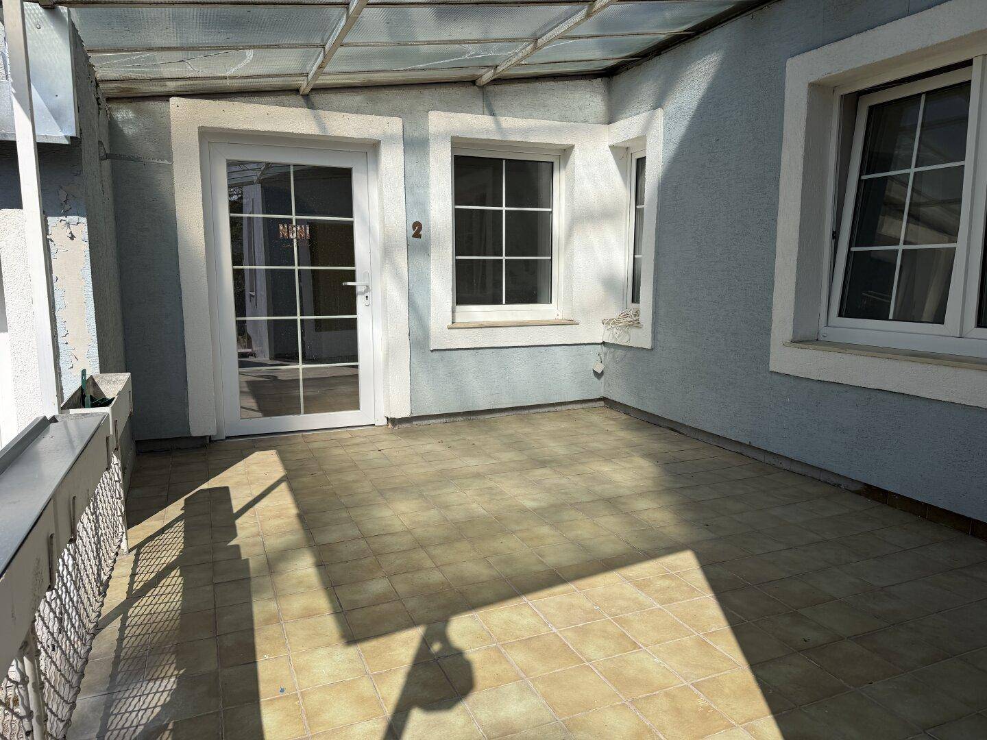 Überdachte Terrasse mit Fliesenboden und Fenstern, die zum Haus führen.