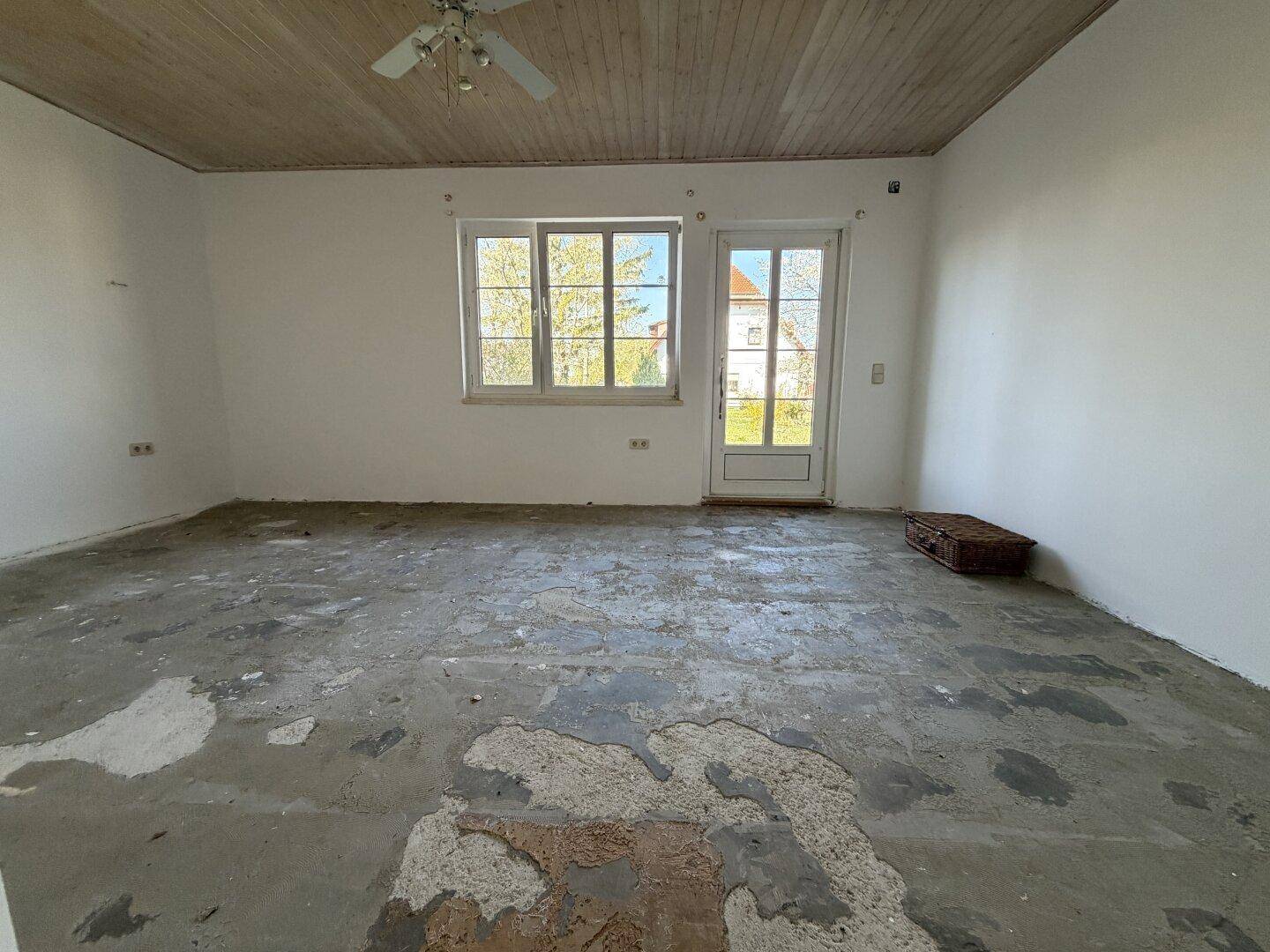 Leerer Raum mit Betonboden, weiß gestrichenen Wänden und einer Holzbalkendecke, bereit für Renovierung.