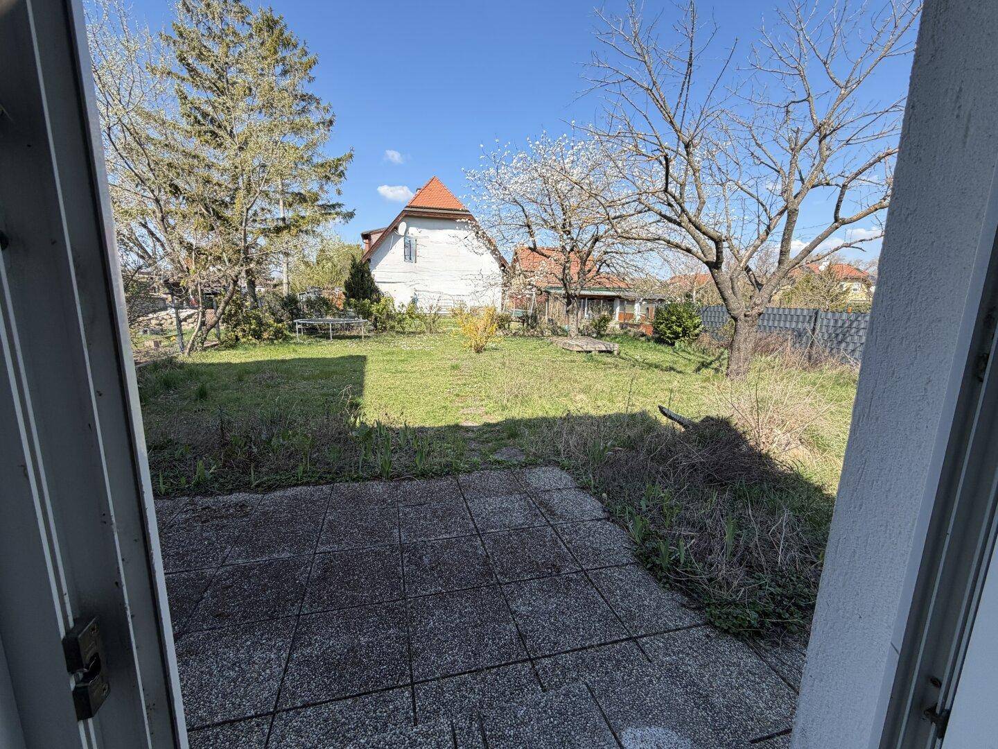 Blick von der Terrasse auf einen weitläufigen Garten mit Bäumen und einem Haus im Hintergrund unter blauem Himmel.