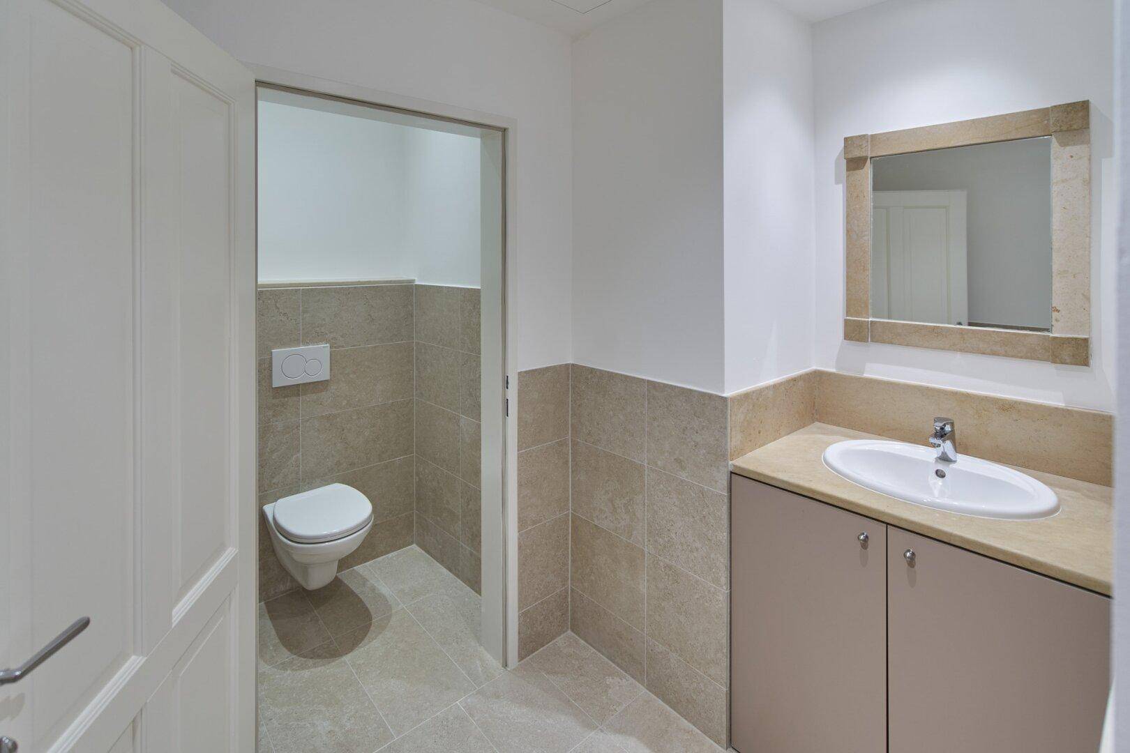 Modernes WC mit Wand-WC, Waschbecken und hellen Fliesen an Wänden und Boden.