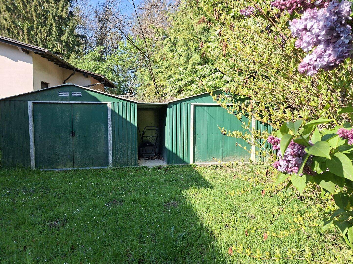 Zwei grüne Gartenschuppen auf einer Rasenfläche, umgeben von Büschen.