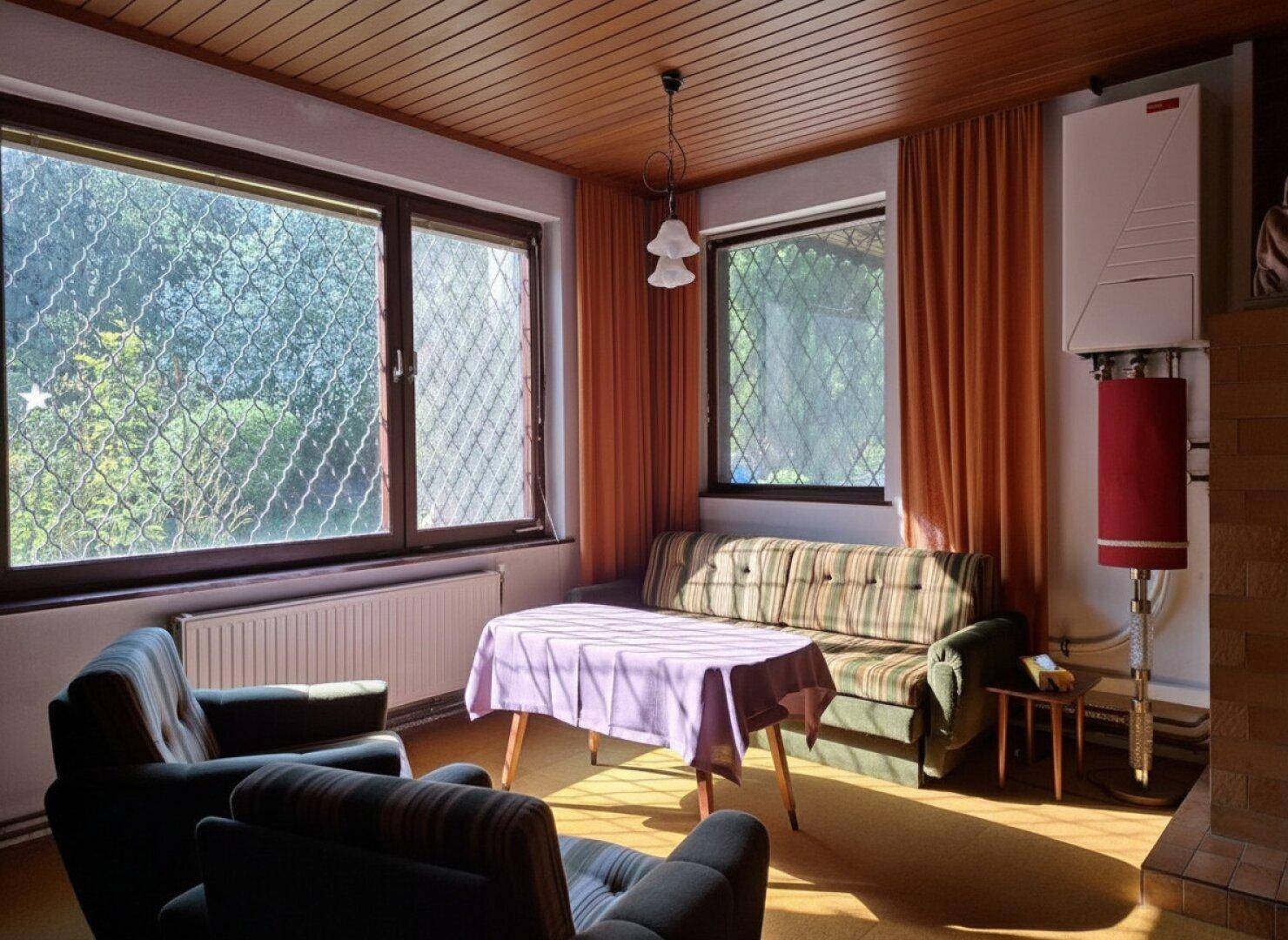 Wohnzimmer mit Heizung, Holzdecke und Blick ins Grüne durch große Fenster.