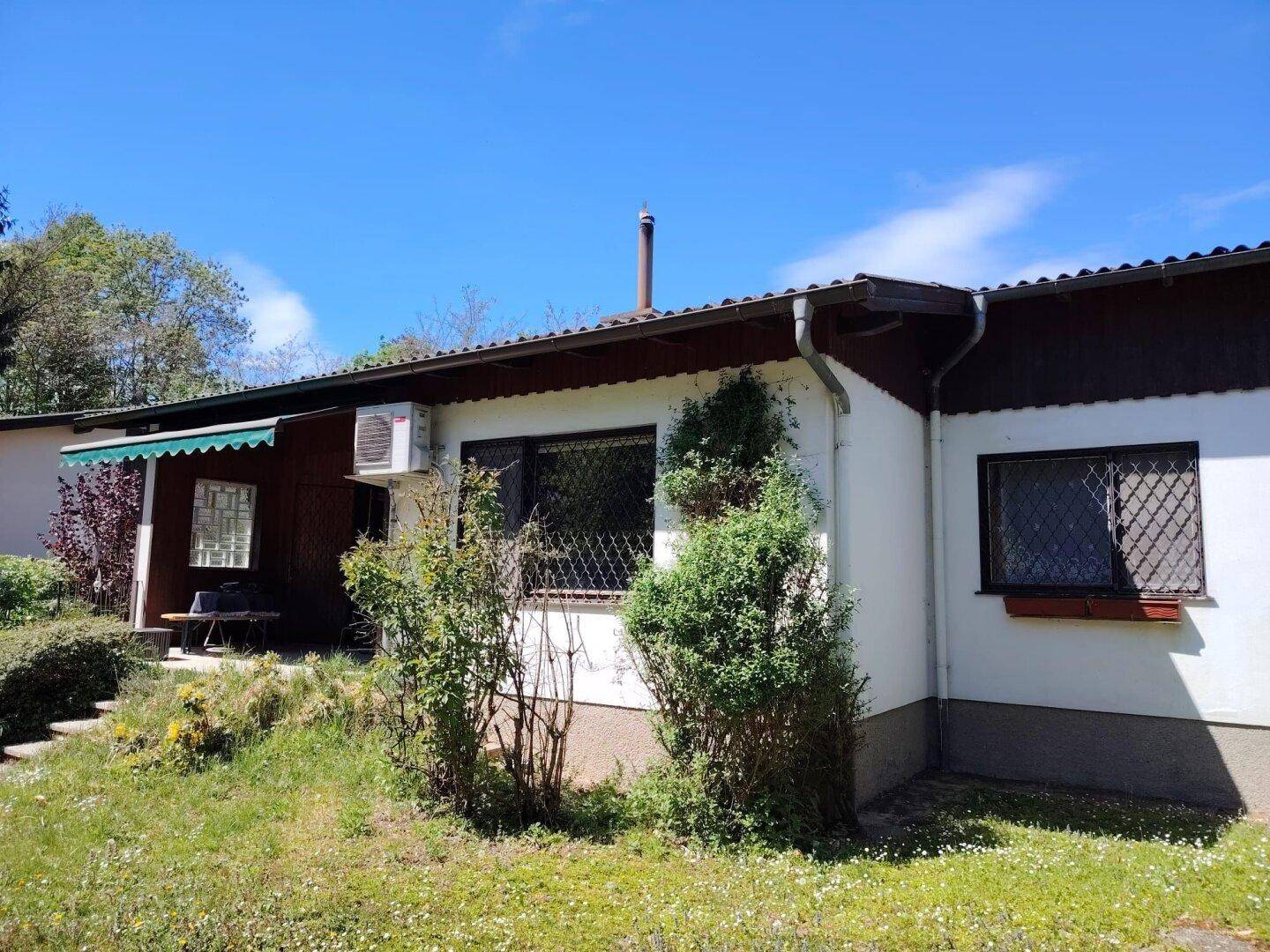 Hausfassade mit kleiner Terrasse und Gartenbereich unter blauem Himmel.