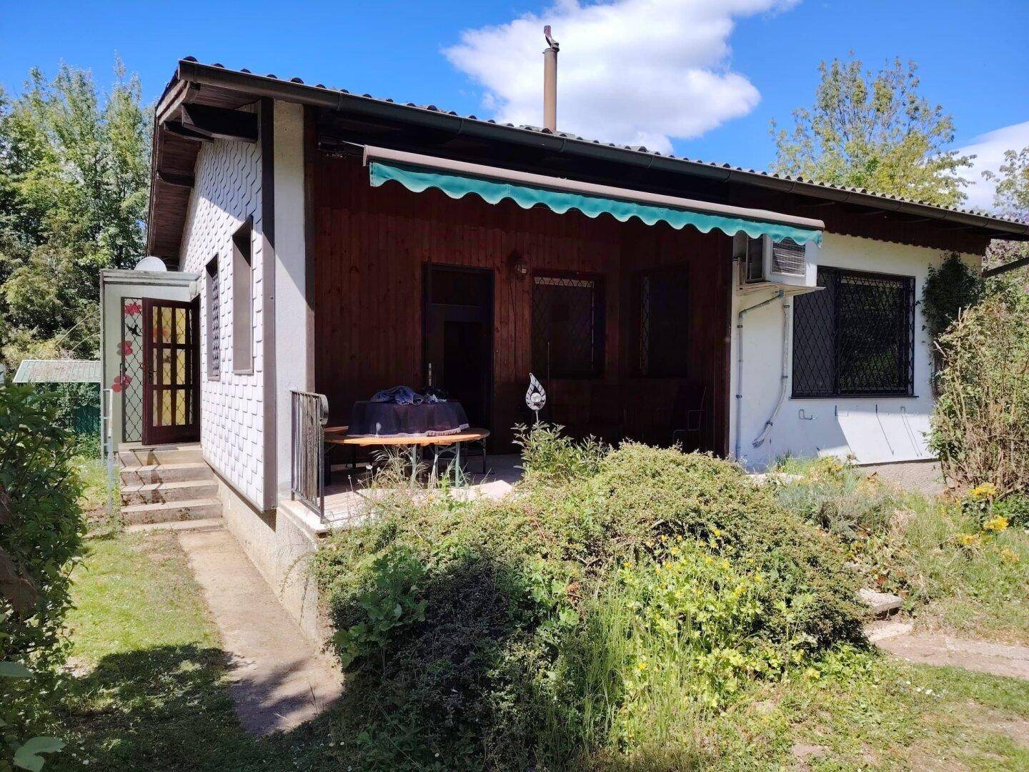 Überdachte Terrasse mit Markise und Holzwänden, umgeben von üppigem Grün.