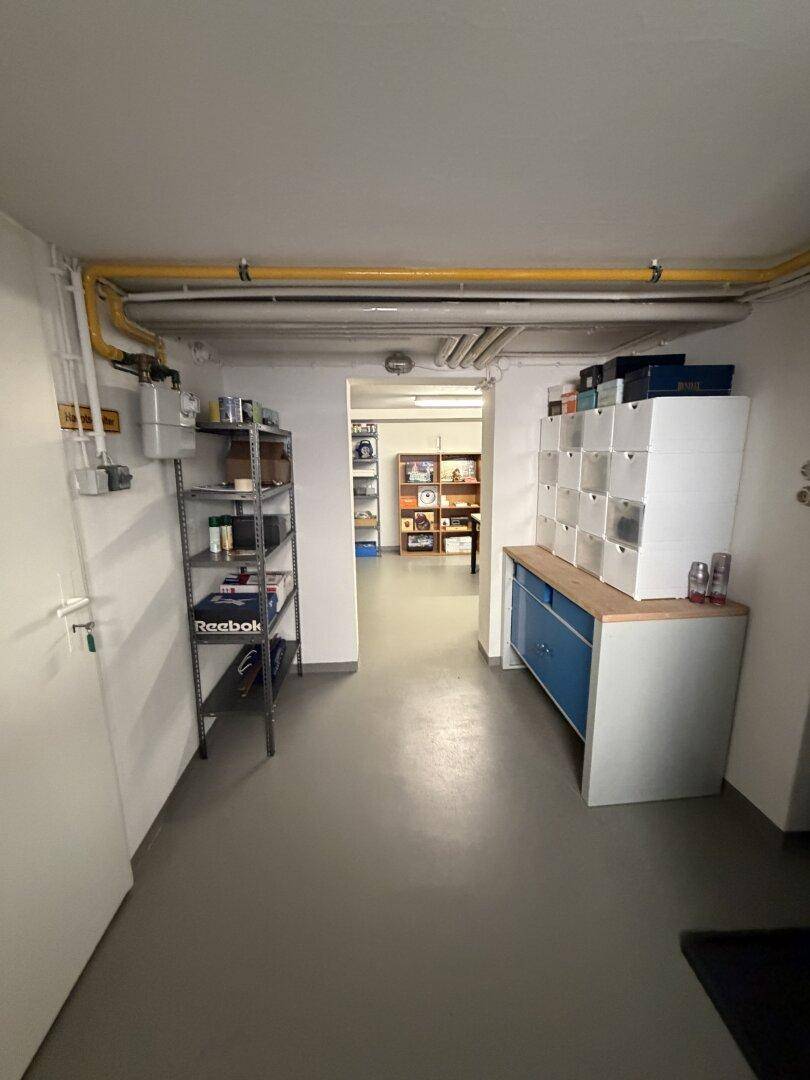 Kellerflur mit Regalen, Schränken und sichtbaren Versorgungsleitungen an der Decke.