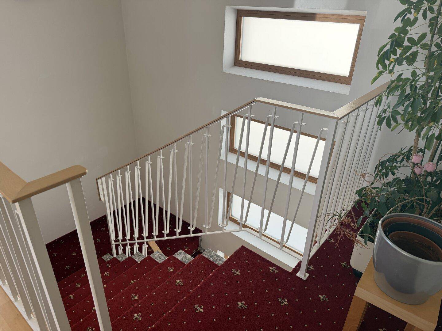 Treppenhaus mit rotem Teppichboden, weißem Geländer und Fenstern für Tageslicht.