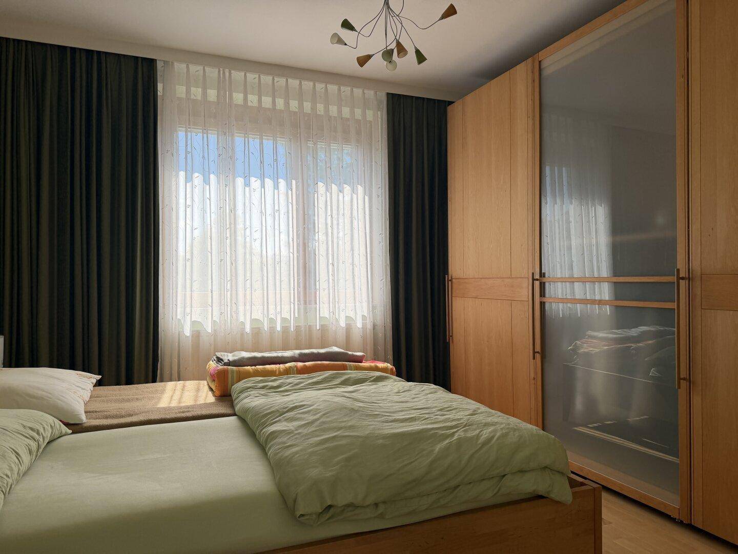 Schlafzimmer mit zwei Einzelbetten, Holzschrank und großem Fenster mit viel Tageslicht.